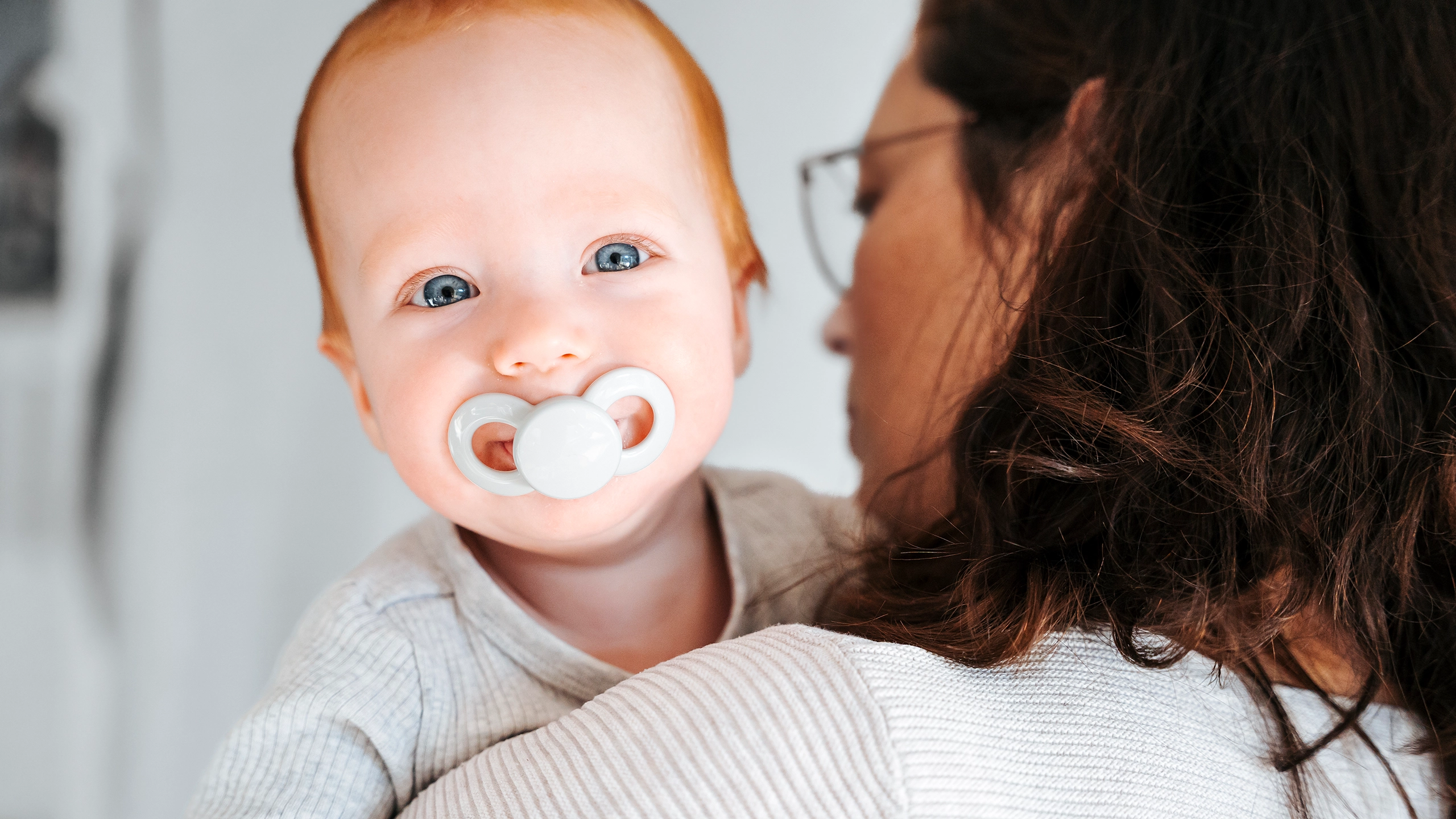 Baby mit weißem Schnuller blickt über die Schulter seiner Mutter in die Kamera